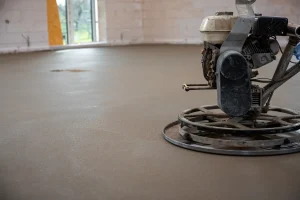 Ein frisch verlegter und geglätteter Estrichboden in einem Rohbau. Im Vordergrund ist eine Schleifmaschine zu sehen, die für die Oberflächenbearbeitung verwendet wird. Die Fläche befindet sich im Zustand vor der CM-Messung zur Bestimmung der Restfeuchte.