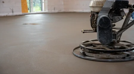 Ein frisch verlegter und geglätteter Estrichboden in einem Rohbau. Im Vordergrund ist eine Schleifmaschine zu sehen, die für die Oberflächenbearbeitung verwendet wird. Die Fläche befindet sich im Zustand vor der CM-Messung zur Bestimmung der Restfeuchte.