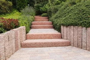 Ein Gartenweg führt hangaufwärts über mehrere Treppenstufen aus rötlichem Naturstein. Die Stufen sind von üppigem, gepflegtem Grün eingefasst. Links und rechts sichern Mauerelemente aus dem gleichen Steinmaterial den Hang. Die Szene wirkt geordnet, naturnah und einladend.