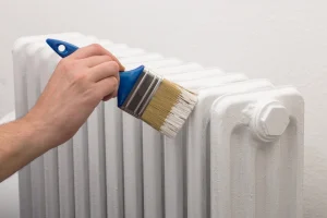 Hand mit Pinsel streicht Rippenheizkörper mit weißer Farbe.