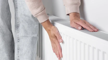 Eine Person trägt beide Hände an einen weißen Heizkörper, um dessen Temperatur zu prüfen. Die Heizung scheint dauerhaft warm zu sein und lässt sich nicht ausschalten. Im Bildausschnitt sind Arme, Hände und ein Teil der Kleidung sichtbar.