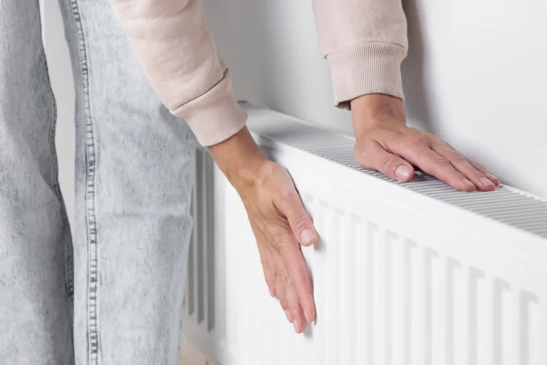 Eine Person trägt beide Hände an einen weißen Heizkörper, um dessen Temperatur zu prüfen. Die Heizung scheint dauerhaft warm zu sein und lässt sich nicht ausschalten. Im Bildausschnitt sind Arme, Hände und ein Teil der Kleidung sichtbar.