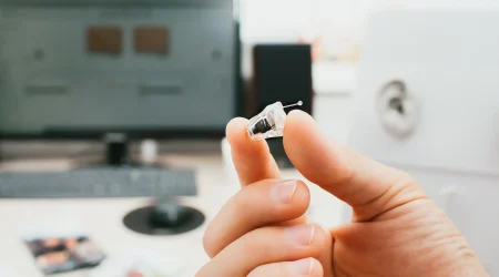 Eine Hand hält ein transparentes ICC Hörgerät zwischen Daumen und Zeigefinger. Das ICC Hörgerät besitzt eine kleine Zugöse und feine elektronische Bauteile im Inneren.