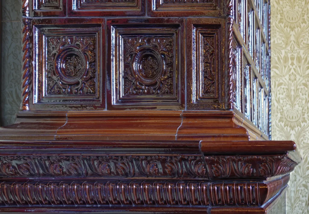 Nahaufnahme eines antiken Kachelofens mit glasierten, braunroten Keramikkacheln und aufwendigen Ornamenten. Die reliefartigen Verzierungen zeigen florale und geometrische Muster. Der Ofen steht vor einer Tapete mit historischem Dekor.
