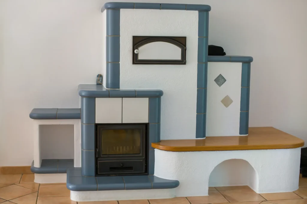 Ein modern gestalteter Kachelofen mit abgerundeten, blau glasierten Kachelrändern und verputzter weißer Oberfläche steht in einem Wohnraum mit Fliesenboden. Der Ofen verfügt über eine integrierte Sitzbank mit Holzauflage sowie über eine geschlossene Feuerraumtür.