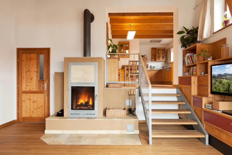 Ein moderner Kachelofen mit verputzter Oberfläche und keramisch umrahmter Feuerraumtür ist in ein erhöhtes Podest im Wohnbereich integriert. Das sichtbare Feuer brennt hinter einer Glastür. Rechts daneben führt eine offene Holztreppe in den höher gelegenen Küchen- und Essbereich.