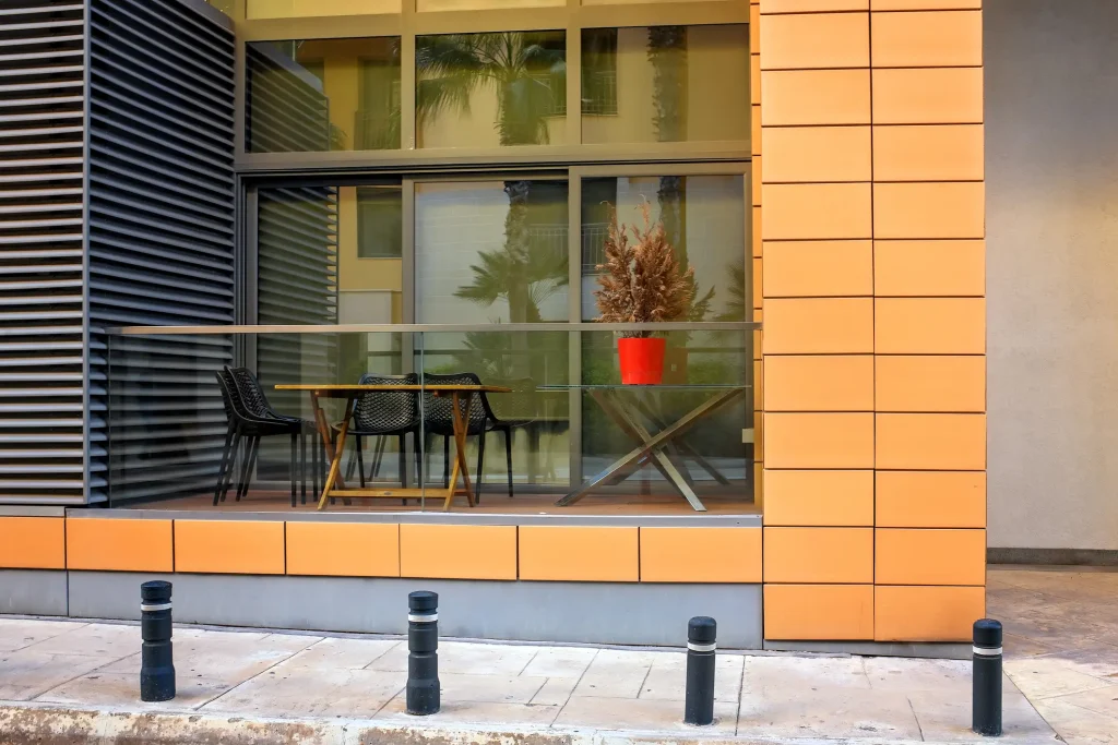 Eine moderne Loggiafläche mit Glasgeländer zeigt einen kleinen Tisch mit zwei schwarzen Stühlen. Auf einem Beistelltisch steht ein roter Pflanztopf mit einer trockenen Zimmerpflanze. Hinter der Loggia befinden sich große Fensterflächen mit Spiegelungen von Palmen und Gebäudefassaden.