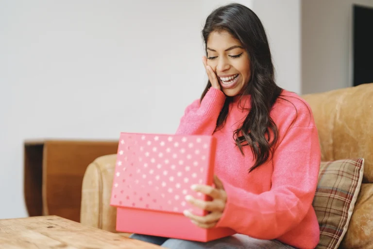Frau in pinkem Pullover öffnet ein Geschenk und freut sich darüber.