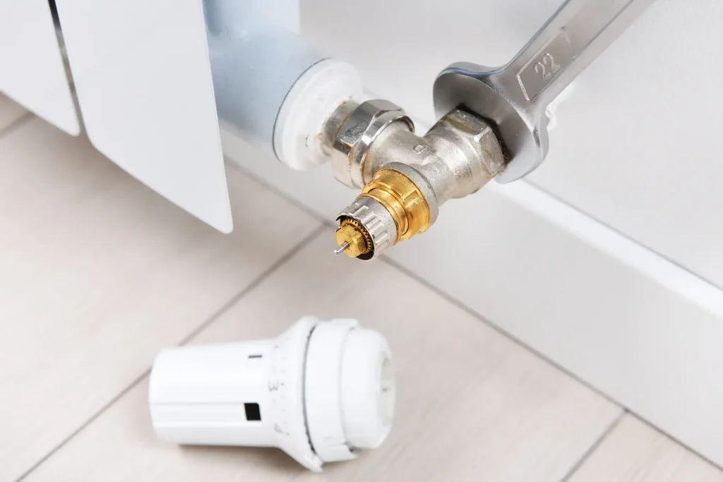 Ein abgenommener Thermostatkopf liegt auf dem Boden vor einem Heizkörper. Am geöffneten Heizkörperventil ist der Ventilstift zu sehen. Ein Maulschlüssel mit der Schlüsselweite 22 sitzt am Verschraubungsgewinde des Ventils.