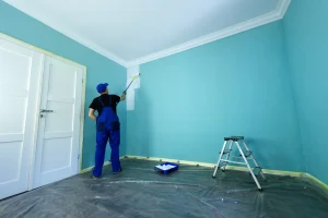 Ein Handwerker in blauer Latzhose streicht mit einer Farbrolle eine türkisfarbene Wand weiß. Der Boden ist sorgfältig mit Schutzfolie ausgelegt, die weißen Türrahmen sind mit Kreppband abgeklebt. Eine Farbwanne und eine Trittleiter stehen griffbereit.