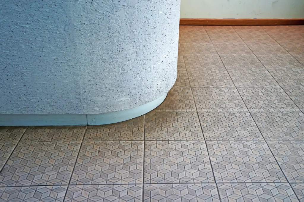 Ein Sockel aus Buntsteinputz mit einer feinen, dunklen Gesteinskörnung bildet eine abgerundete Kante und sitzt auf einem Fliesenboden mit braunem, geometrischem Sternenmuster.