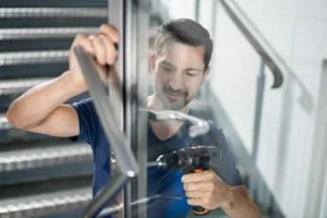 Ein Handwerker montiert fachgerecht den Türgriff an einer transparenten Glastür mit einem Akkuschrauber in einem Treppenhaus.