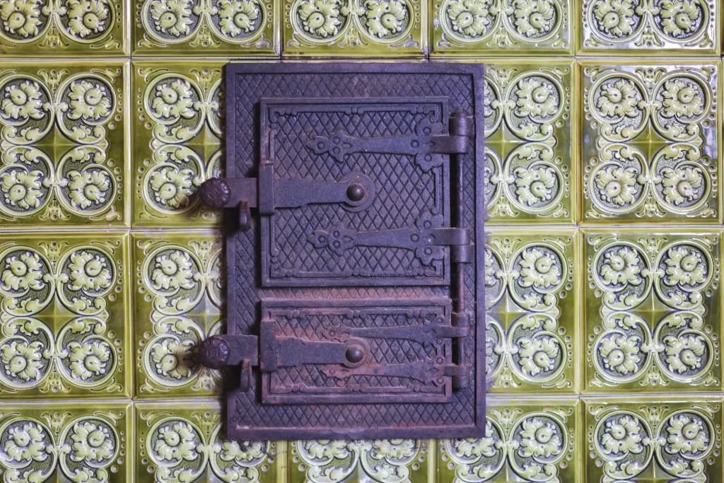 Historische Kachelofen-Tür aus dunklem Gusseisen mit kunstvollen Verzierungen eingebettet in eine Wand aus olivgrünen verzierten Keramikkacheln.
