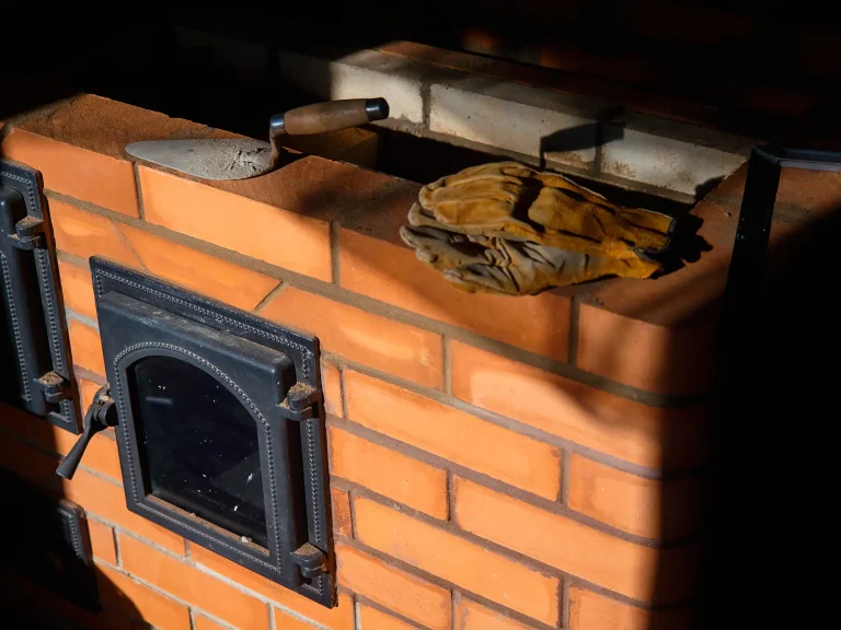 Einbau eines Kachelofens aus roten Schamottsteinen mit gusseisernen Ofentüren, daneben liegen Maurerkelle und Arbeitshandschuhe.