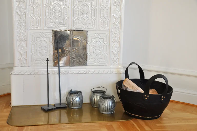 Historischer weiß glasierter Kachelofen mit verzierten Ofenkacheln und geöffneter Reinigungsklappe, davor Kaminbesteck, Zinkgefäße und ein Korb mit Brennholz.