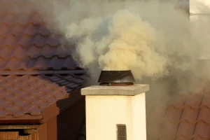 Ein Kaminbrand auf einem Hausdach mit braunen Dachziegeln. Dichter, heller Rauch quillt unter hohem Druck aus der schwarzen Mündung eines gemauerten, hell verputzten Schornsteins. Der Rauch breitet sich über die Dachfläche aus und verdeckt teilweise den Hintergrund.