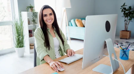 Eine Frau sitzt mit Bluse im Homeoffice vor ihrem Bildschirm. Im Hintergrund ist ein helles modernes Wohnzimmer zu erkennen.