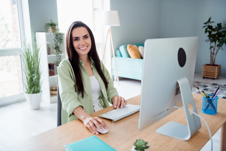 Eine Frau sitzt mit Bluse im Homeoffice vor ihrem Bildschirm. Im Hintergrund ist ein helles modernes Wohnzimmer zu erkennen.