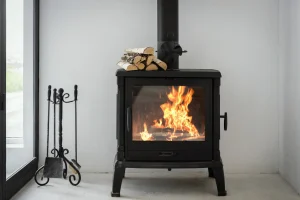 Ein moderner schwarzer Kaminofen mit brennendem Holzfeuer hinter einer Glasscheibe, daneben steht ein Kaminbesteck vor einer weißen Wand.