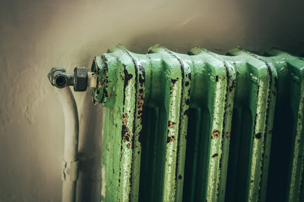 Nahaufnahme eines alten, grün lackierten Gliederheizkörpers aus Gusseisen, der deutliche Roststellen, abplatzende Farbe und Gebrauchsspuren an den Rippen zeigt.