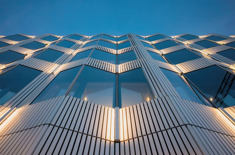 Moderne Glas und Metallfassade mit geometrischen Mustern die bei Einbruch der Dunkelheit beleuchtet wird.