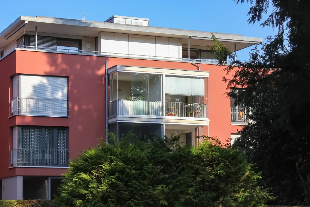 Außenansicht eines modernen mehrstöckigen Wohnhauses mit roter Fassade und einer raumhohen Balkonverglasung, die das Geländer umschließt und als Wetterschutz dient.