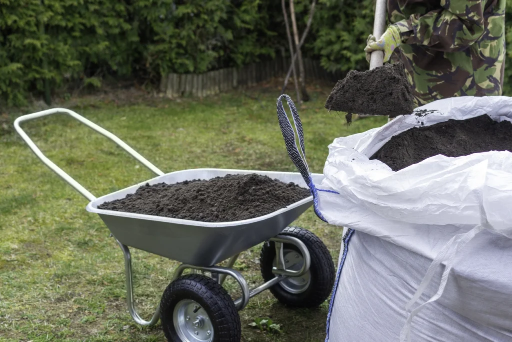 Nahaufnahme einer Person im Camouflage-Anzug, die mit einer Schaufel dunkle Erde aus einem großen weißen Big Bag in eine silberne Schubkarre mit zwei Rädern füllt, die auf einer Rasenfläche steht.