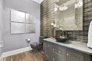 Modernes Gäste-WC mit einer Akzentwand aus Tapete in Grau-Gold-Optik, die direkt über die alten Fliesen tapeziert wurde, kombiniert mit einem dunklen Aufsatzwaschbecken und einem großen Zierspiegel.