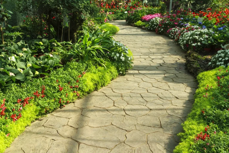 Ein geschwungener Gartenweg aus hellen Natursteinen in Polygonaloptik, der durch eine üppig blühende Gartenanlage mit roten, weißen und rosa Blumenbeeten führt.