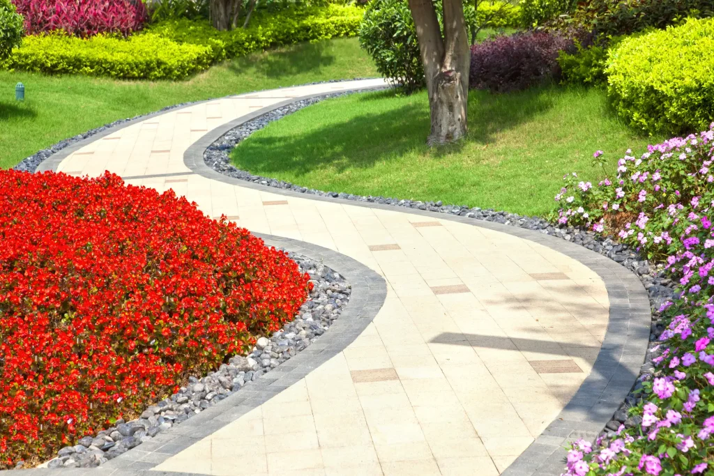 Gartenweg aus großformatigen, beigefarbenen Platten, die bündig mit einer dunkelgrauen Steinkante eingefasst sind. Direkt daneben verläuft ein Zierstreifen aus hellem Kies und eine Rasenfläche.