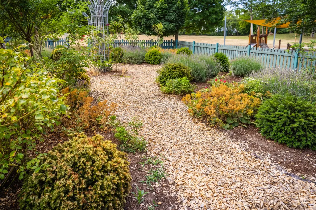 Ein natürlich angelegter Gartenweg aus hellem Rindenmulch oder Holzhackschnitzeln, der durch ein bepflanztes Beet mit Stauden und Sträuchern führt. Im Hintergrund begrenzt ein grüner Holzzaun das Grundstück.