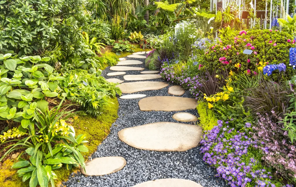 Ein geschwungener Gartenweg aus flachen, ovalen Trittsteinen aus Naturstein, die in einem Bett aus dunklem Edelsplitt verlegt sind und durch eine üppig bepflanzte Gartenanlage mit lila, gelben und pinkfarbenen Blüten führen.