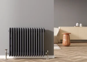 Ein moderner, anthrazitfarbener Gliederheizkörper ist an einer grauen Wand in einem stilvoll eingerichteten Raum mit Fischgrätparkett montiert. Im Hintergrund ist eine helle Kommode sichtbar.
