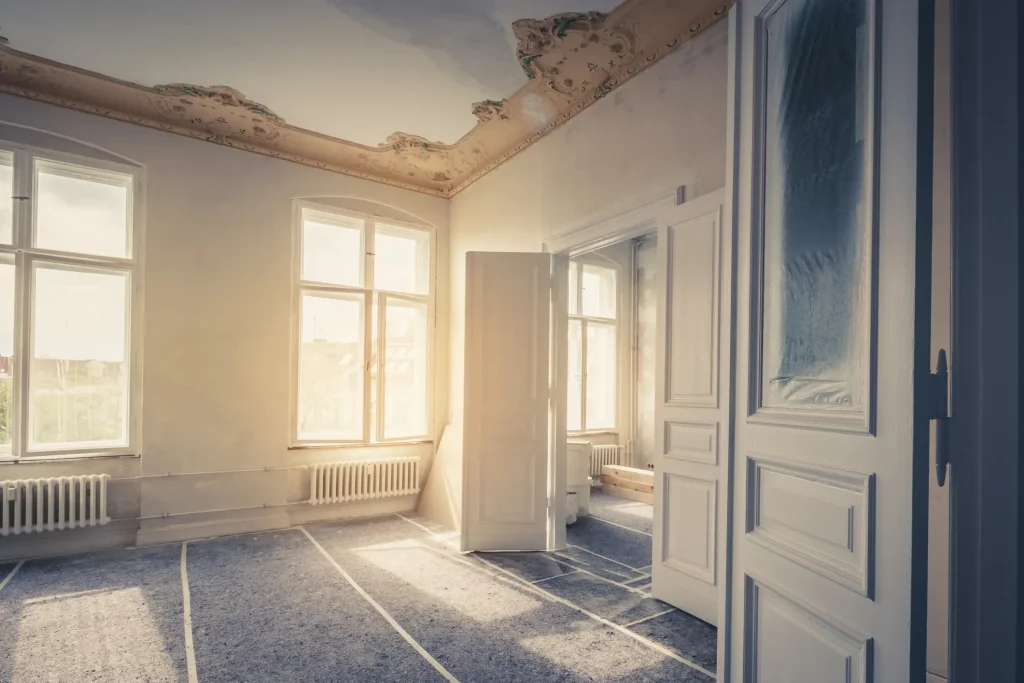 Heller, leerstehender Altbauraum während der Sanierung mit hohen Fenstern, Stuckverzierung an der Decke und klassischen weißen Gliederheizkörpern unter den Fensterbänken.