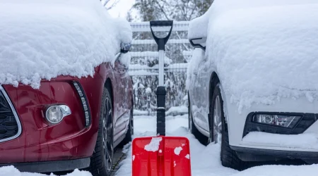 Zwei eingeschneite Autos parken nebeneinander. Zwischen ihnen steht eine rote Schneeschaufel.