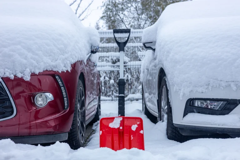 Zwei eingeschneite Autos parken nebeneinander. Zwischen ihnen steht eine rote Schneeschaufel.