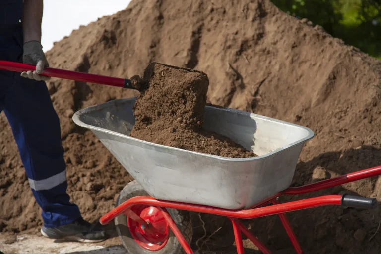Ein Gärtner füllt mit einer Schaufel dunklen, frischen Mutterboden in eine rote Schubkarre. Im Hintergrund ist ein großer Erdhaufen auf einer Baustelle oder in einem Garten zu sehen.