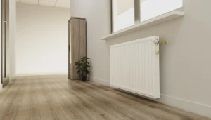 Ein moderner weißer Plattenheizkörper ist unter einem Fenster in einem hellen Raum mit hellem Laminatboden montiert. Im Hintergrund sind ein Holzschrank und eine Zimmerpflanze zu sehen.