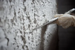Nahaufnahme eines Malers, der mit einer Glättkelle eine strukturierte, weiße Rauputzschicht grob auf eine Wand aufträgt.