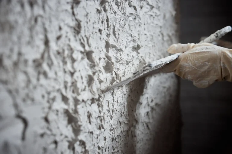 Nahaufnahme eines Malers, der mit einer Glättkelle eine strukturierte, weiße Rauputzschicht grob auf eine Wand aufträgt.