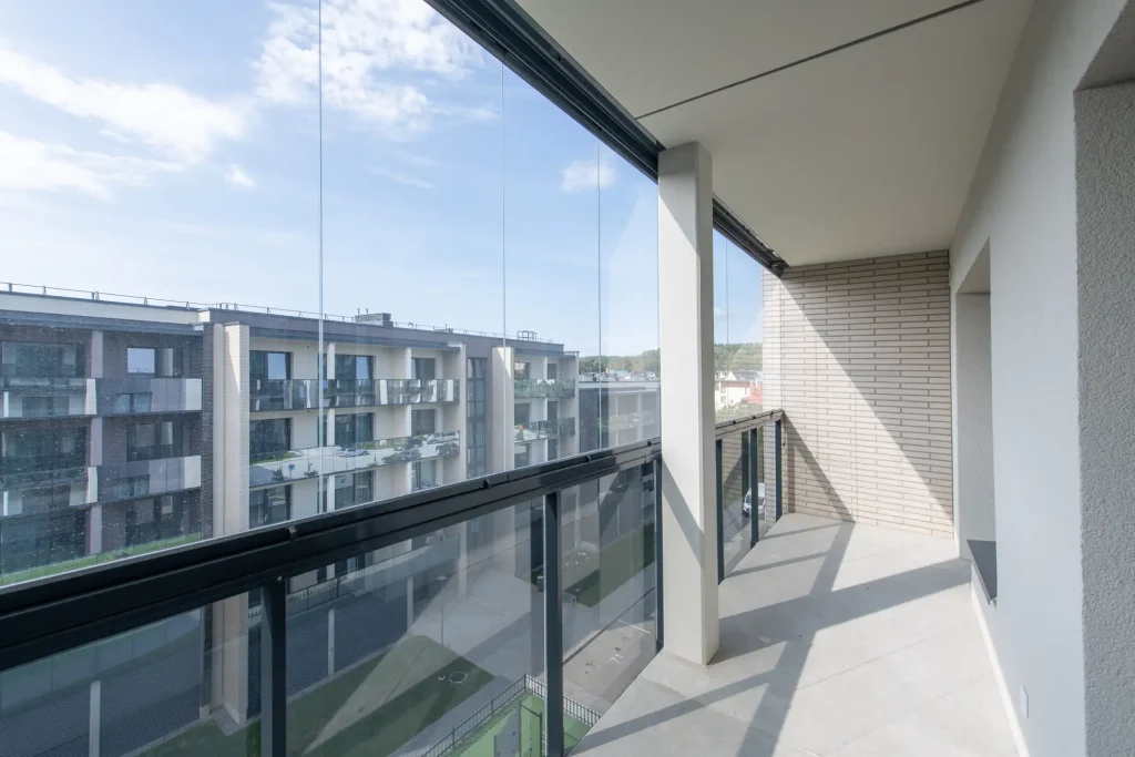 Innenansicht eines modernen, langen Balkons mit einer transparenten Schiebe-Verglasung und einem dunklen Metallgeländer, die einen freien Blick auf gegenüberliegende Apartmenthäuser bietet.