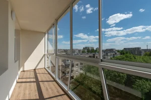 Ein heller, geschlossener Balkon mit einer rahmenlosen Balkonverglasung, die einen weiten Blick über eine Stadtlandschaft und grüne Bäume unter blauem Himmel ermöglicht.