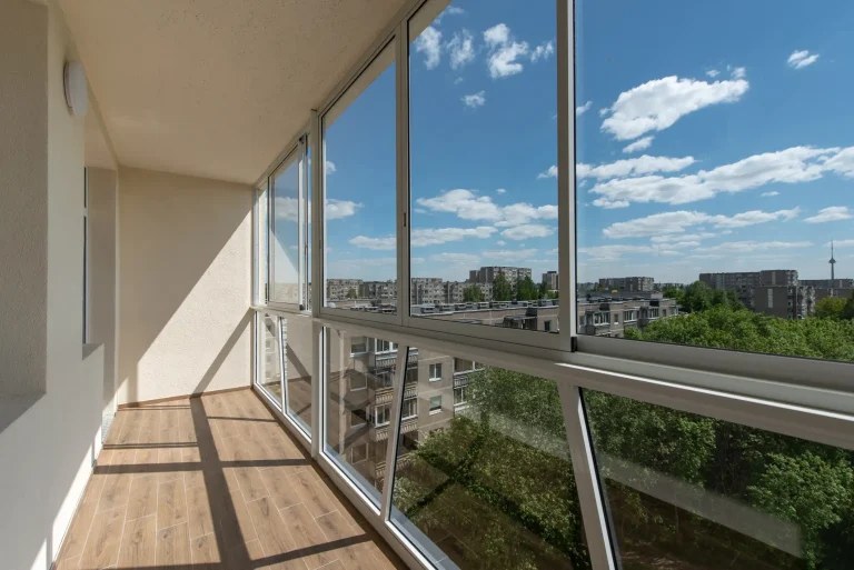 Ein heller, geschlossener Balkon mit einer rahmenlosen Balkonverglasung, die einen weiten Blick über eine Stadtlandschaft und grüne Bäume unter blauem Himmel ermöglicht.