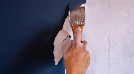 Nahaufnahme einer Hand, die mit einem Metallspachtel alte weiße Farbe von einer dunkelblauen Wand abkratzt, um den Untergrund für eine Neugestaltung vorzubereiten.