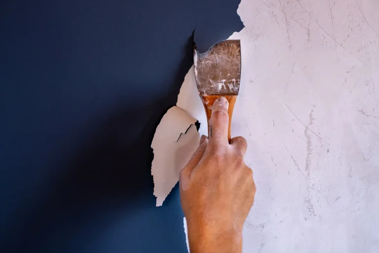 Nahaufnahme einer Hand, die mit einem Metallspachtel alte weiße Farbe von einer dunkelblauen Wand abkratzt, um den Untergrund für eine Neugestaltung vorzubereiten.