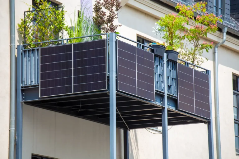 Ein moderner Anbaubalkon aus einer anthrazitfarbenen Metallkonstruktion, der auf Stützen vor der Hausfassade steht. An der Brüstung sind drei Photovoltaik-Module eines Balkonkraftwerks als Sichtschutz montiert.