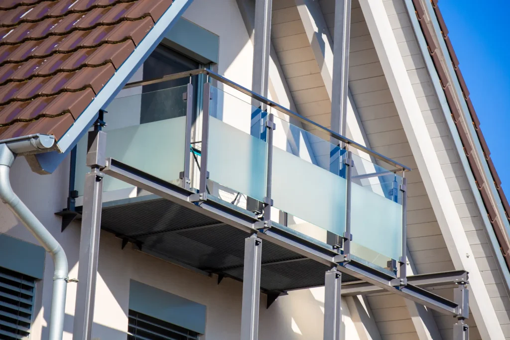 Nachträglich angebauter Anbaubalkon aus einer feuerverzinkten Stahlkonstruktion mit Stützen, montiert an einem Einfamilienhaus. Die Brüstung besteht aus Edelstahlpfosten und satiniertem Sichtschutzglas unter einem Giebeldach.