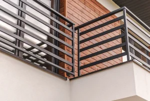 Nahaufnahme eines modernen Balkons mit einer minimalistischen, schwarzen Metallbrüstung aus horizontalen Streben vor einer Fassade mit rötlicher Holzverkleidung und großen Glaselementen.