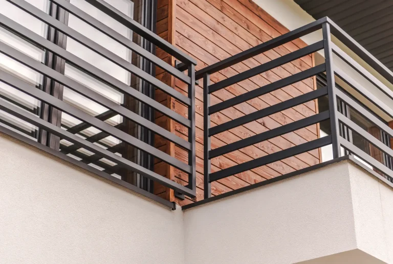 Nahaufnahme eines modernen Balkons mit einer minimalistischen, schwarzen Metallbrüstung aus horizontalen Streben vor einer Fassade mit rötlicher Holzverkleidung und großen Glaselementen.