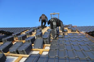 Zwei Dachdecker bei der Verlegung von anthrazitfarbenen Dachziegeln auf einer Holz-Dachkonstruktion unter blauem Himmel.